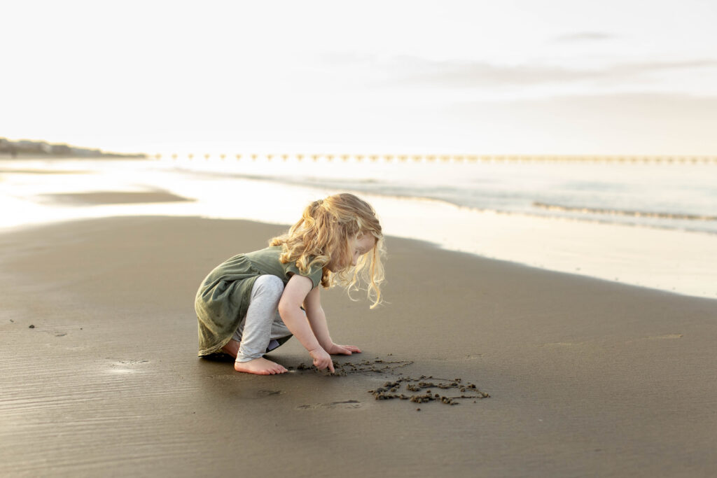 A young girl plays in the wet sand close to the ocean as she draws with her finger during a playful adventurous family photography session during the golden hour in virginia beach