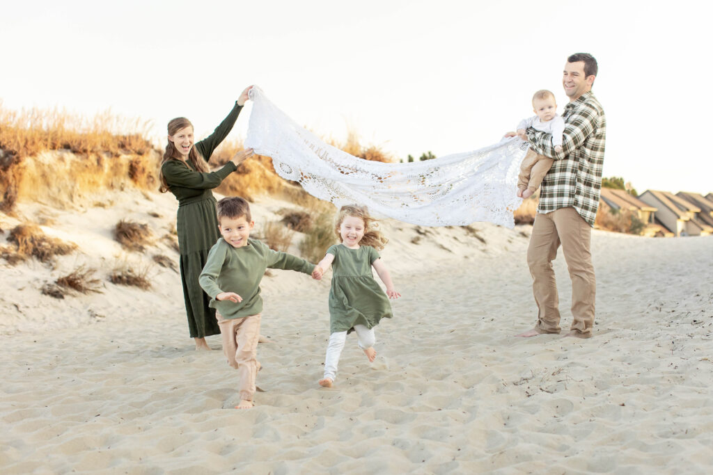 young virginia beach family dressed in neutral colors plays together on the dunes as their children hold hands and run under a blanket laughing together joyfully