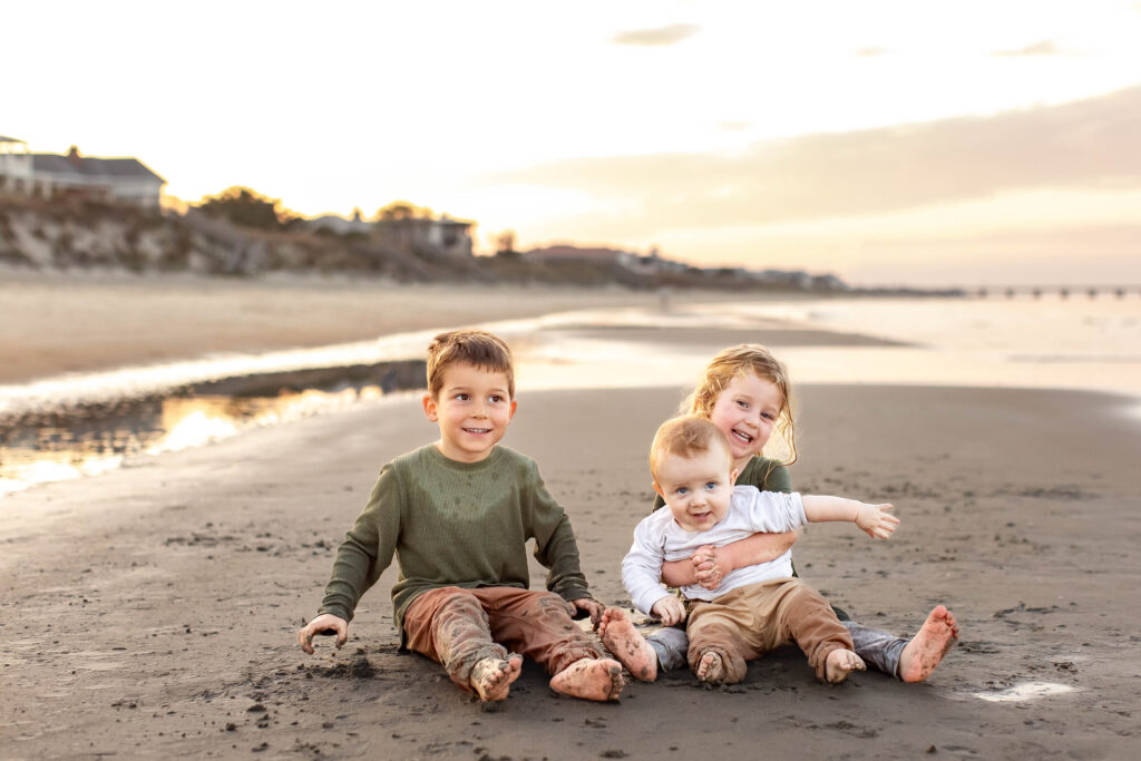 three young siblings sit in the sand together laughing during the golden hour as the sun sets behind them during a family photography session in virginia