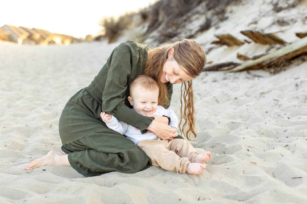 A mother dressed in a long green flowing dressed crouches down on the sand snuggling her baby close as she tickles and hugs him while he laughs as the sun sets behind them on the dunes in virginia beach