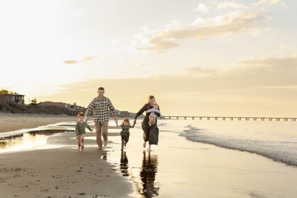 A young family runs together in the shore break, laughing and holding hands during a joyful Virginia Beach family photography session.