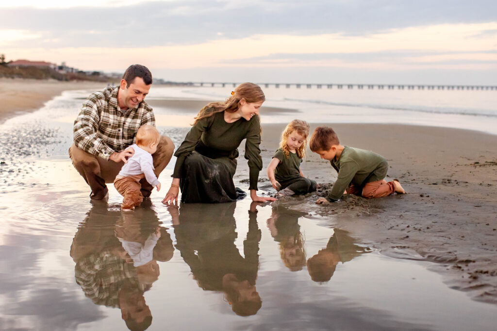 A young family of five dressed in a greens and brown neutral color combination play together in a tidal pool as the sun sets behind them on the Chesapeake bay in Virginia Beach Virginia.  The mom watches her two toddlers build mud castles as the dad laughs holding his baby as he attempts to stand in the shallow water next to them.  