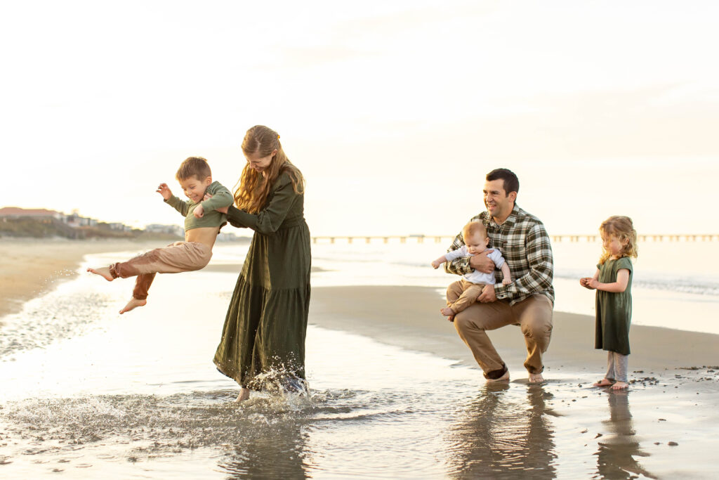 A young family of five plays joyfully and candidly on the beach in Virginia Beach.  The mother dressed in a long flowing green dress is swinging her son from side to side in the water making splashes as her young daughter giggles on the side and her husband cuddles their baby beside her