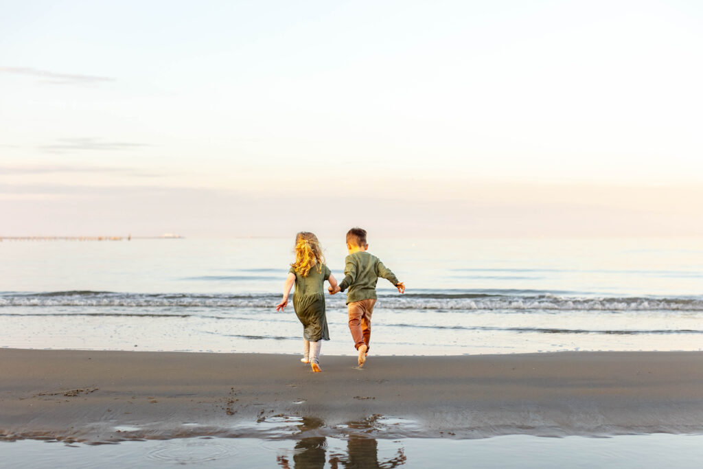 A young brother and sister run and splash joyfully in the ocean holding hands as the sun sets in Virginia Beach