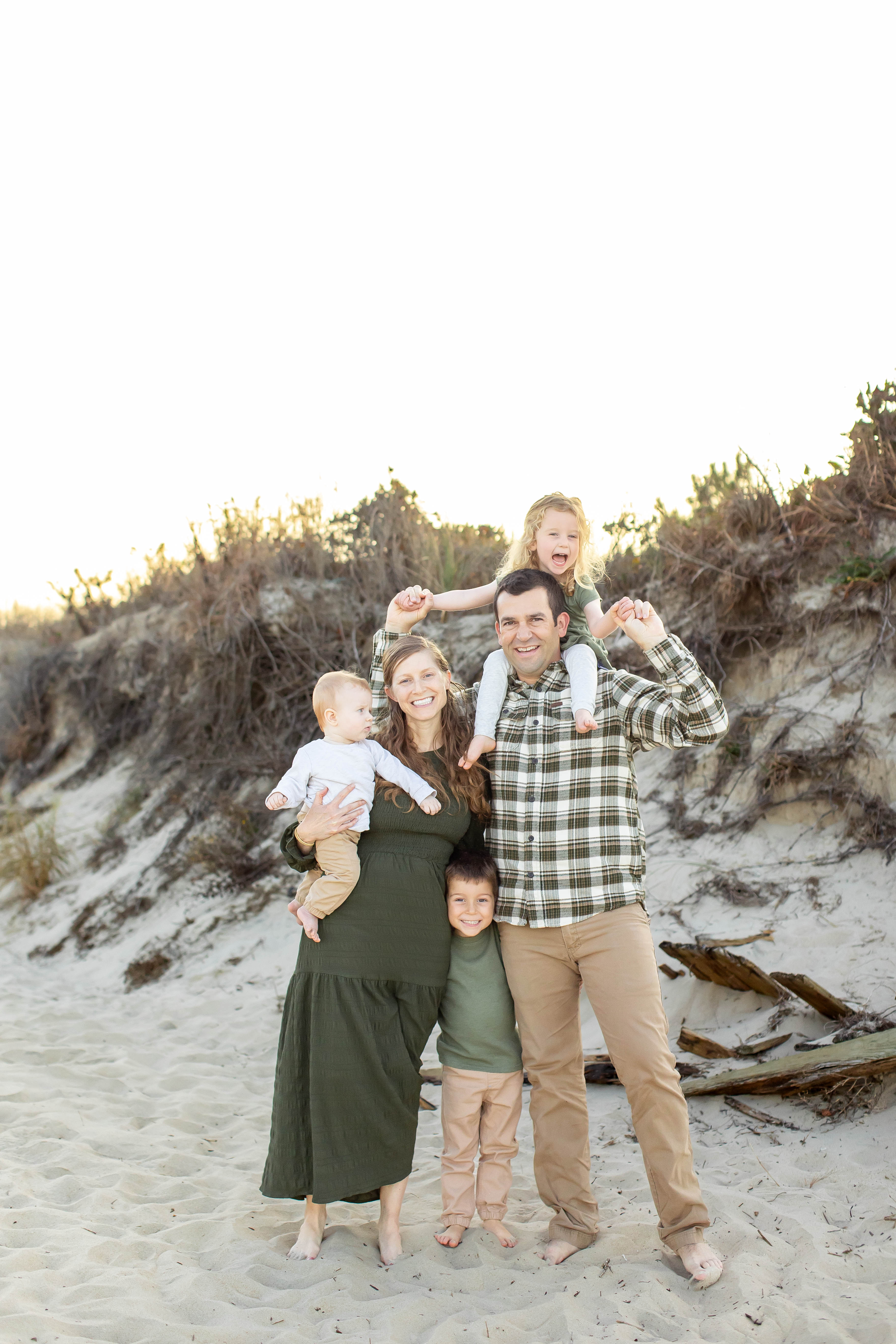 Beautiful young family dressed in green and brown neutrals laugh and play together during a family photo session in Virginia Beach.  The dad holds their tiny daughter on his shoulders as she laughs gleefully while mom holds their baby and their son peeks out from behind mom and dad's legs laughing with a mischievious smile